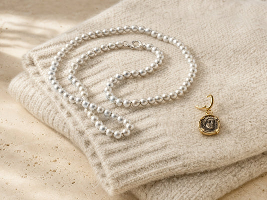 Matte silver bead necklace and a gold-set antique coin charm laid on a folded oatmeal knit jumper - Golden Horn Jewellery